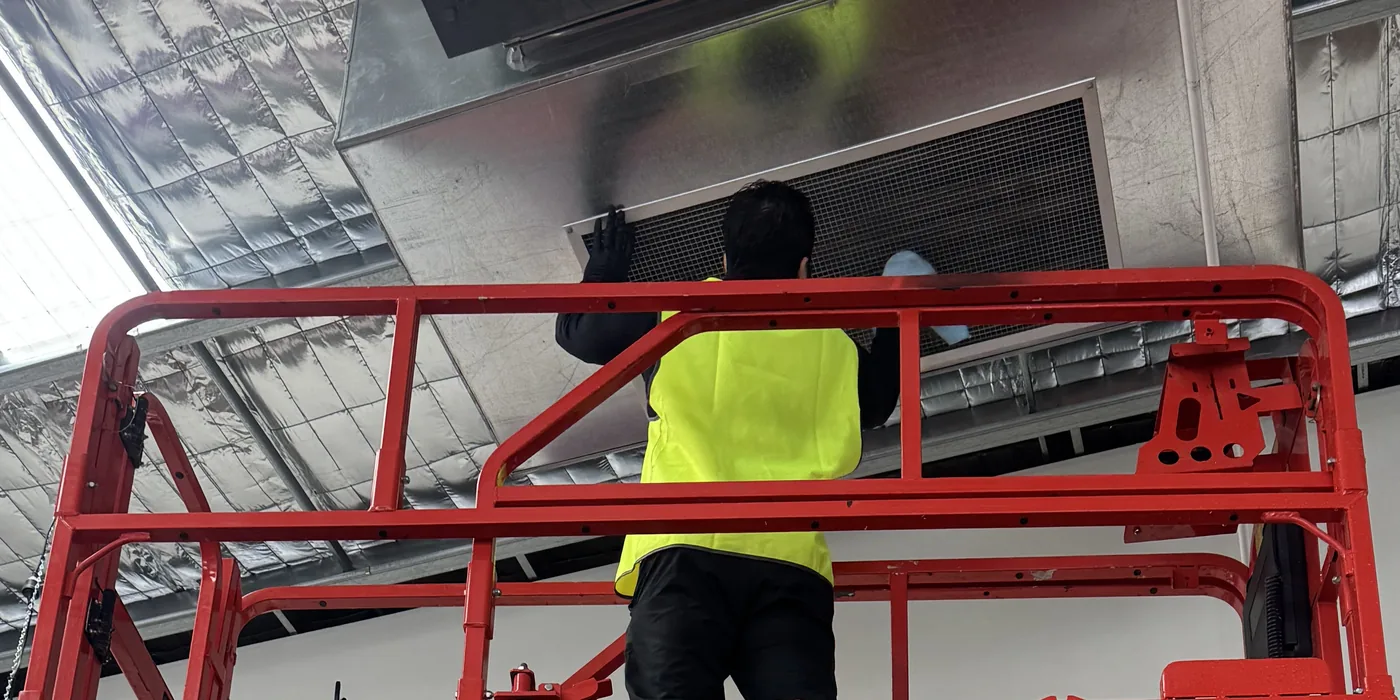A worker on a mobile work platform at an air vent high in the gym roof space during the clean