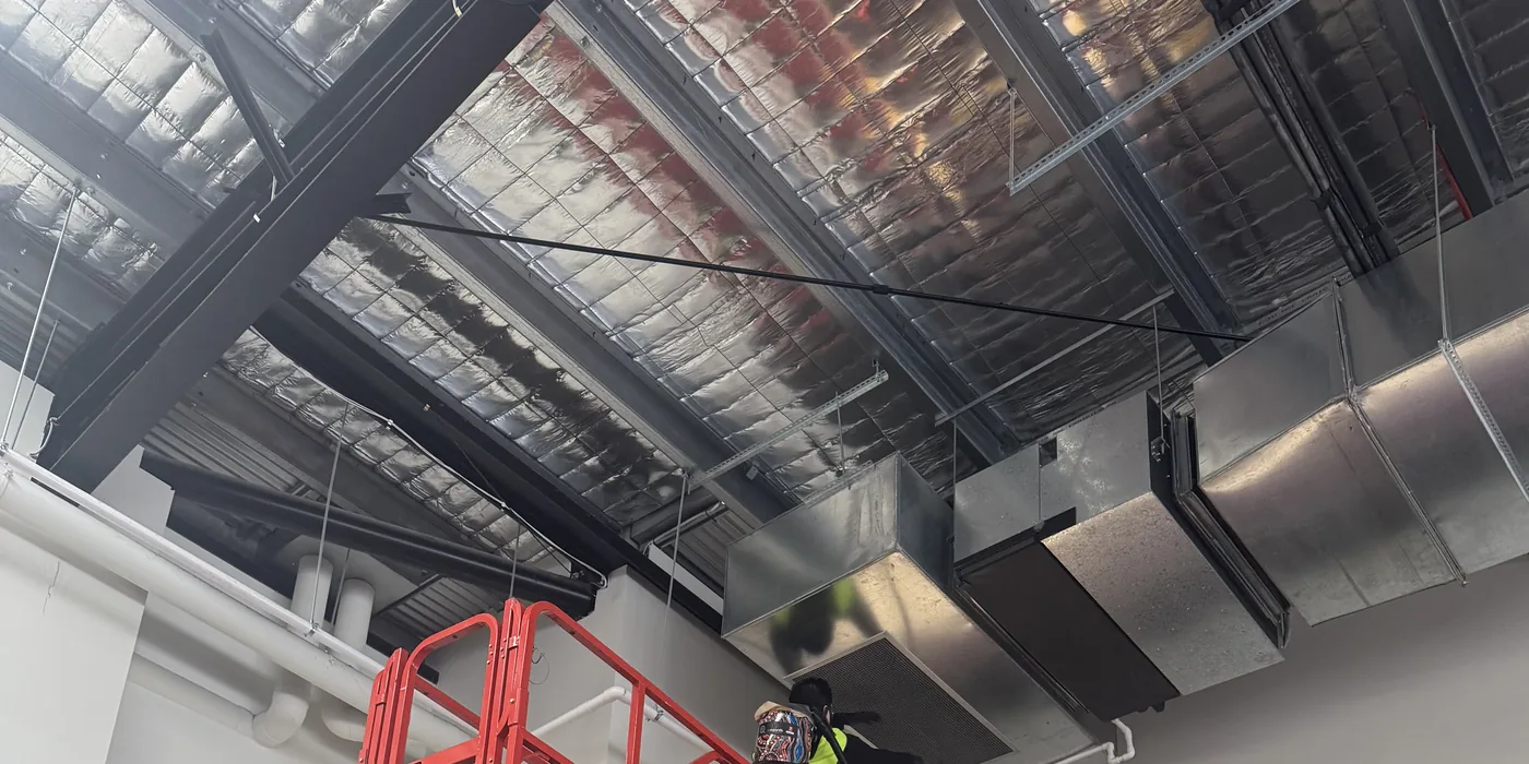A worker in a high visibility vest on a work platform holding a vacuum hose to a ceiling vent grille, with ductwork and roof insulation above