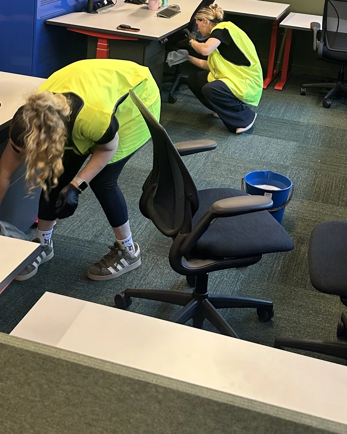 Two cleaners in high visibility vests wiping down desk pedestals and chairs at floor level in an open plan office