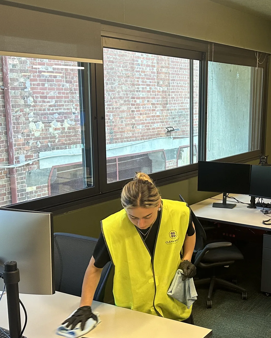 A cleaner in a Clean My Biz vest wiping down a desk in front of the office windows