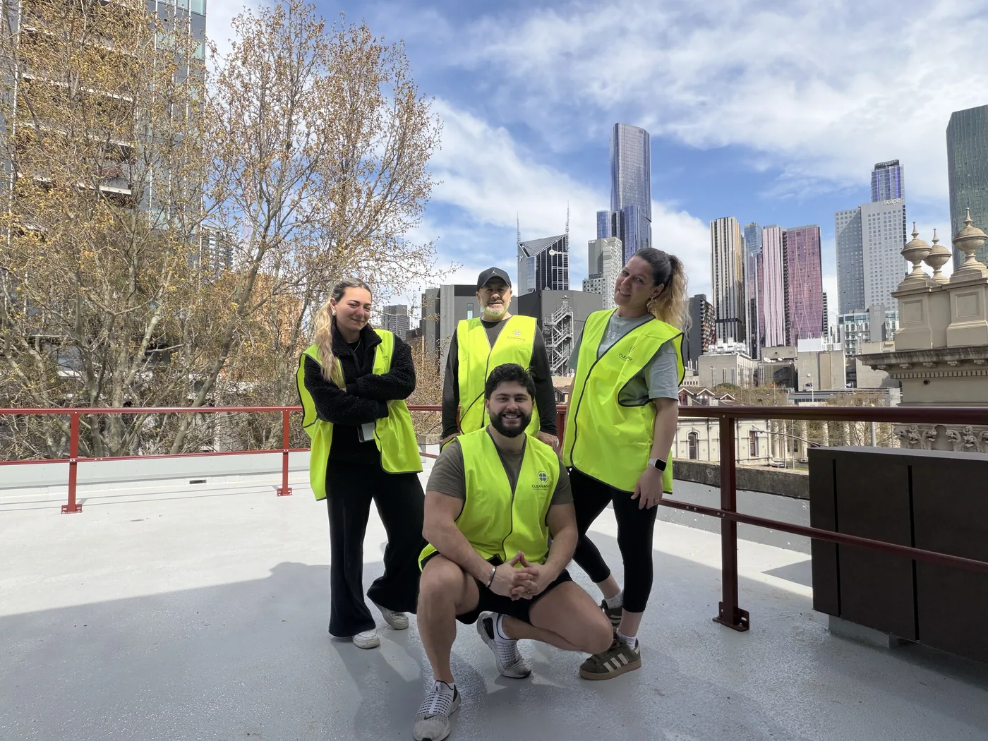 Four Clean My Biz crew members in high visibility vests on a rooftop with the Melbourne skyline behind them