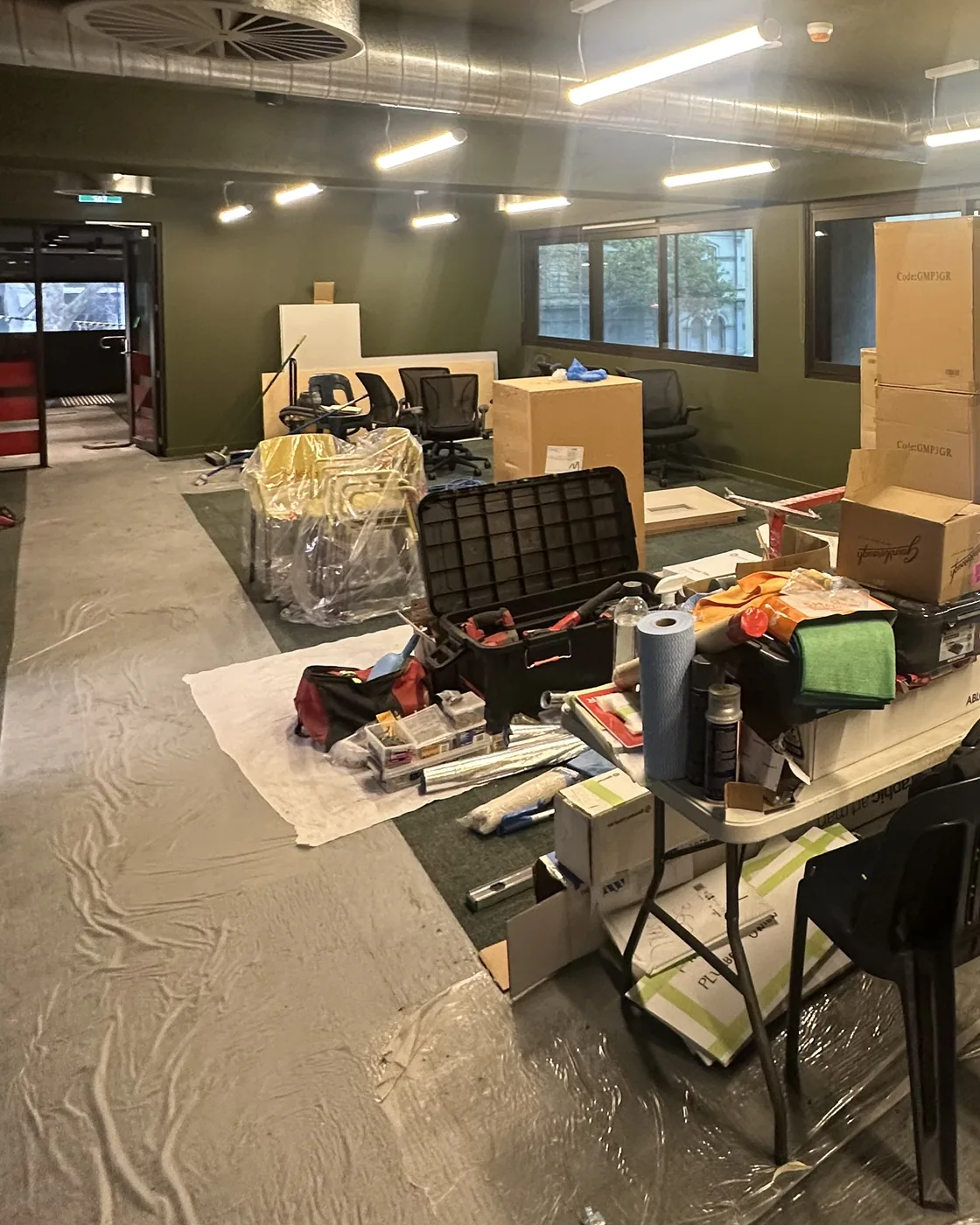 An office floor at the start of the job, with plastic floor protection down, stacked boxes, an open toolbox and a folding table covered in supplies