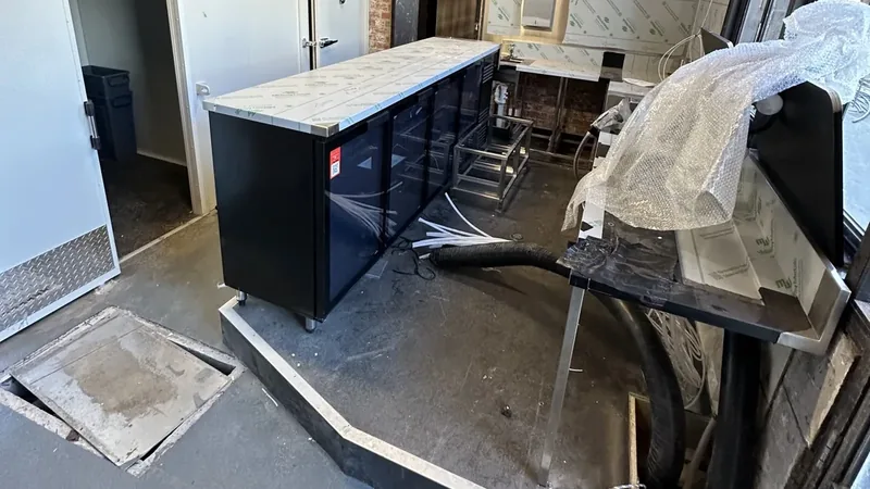 A venue bar fridge still under protective film during a fit-out, with bubble wrap, a loose hose and debris across the floor