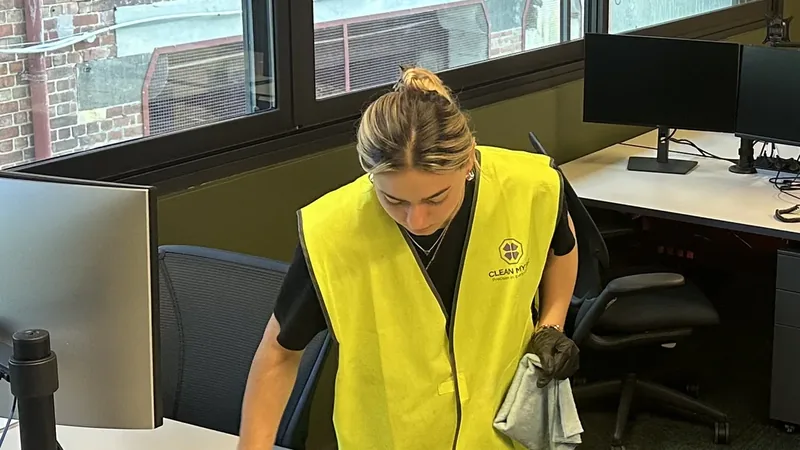 A Clean My Biz cleaner in a high visibility vest wiping down a desk in an office, with the windows and a neighbouring brick wall behind her