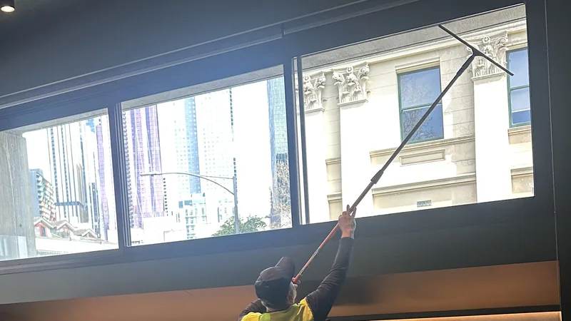 A cleaner in a hi-vis vest, back to the camera, reaching a long-handled squeegee to the top of a tall kitchenette window