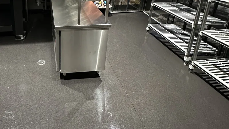 A commercial kitchen with a stainless bench unit, empty shelving racks and a dark polished floor